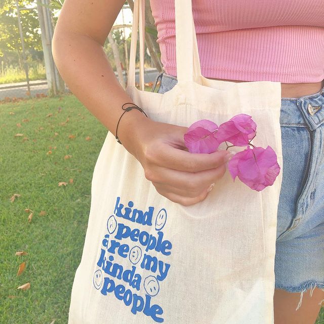 look-verao-com-ecobag Foto do look de uma menina em um parque. Ela usa um top cropped rosa claro, shorts jeans, ecobag com estampa azul e segura uma flor na mão direita.