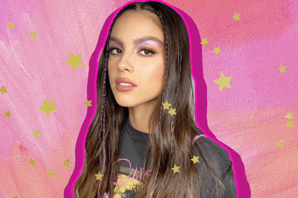 Montagem com foto da Olivia Rodrigo em fundo rosa e estrelinhas douradas. A cantora está olhando para a câmera, de cabelo solto com trancinhas na franja e expressão facial séria, usando uma camiseta cinza.