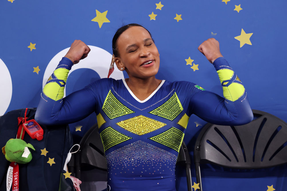 Rebeca Andrade celebrando conquista em Tóquio com os braços em muque. Ela veste um collant azul e amarelo.