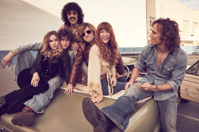 Daisy Jones and The Six – First Look Daisy Jones and The Six; elenco caracterizado como personagens da série reunidos enquanto posam para foto sentados e sorrindo ao ar livre durante o dia
