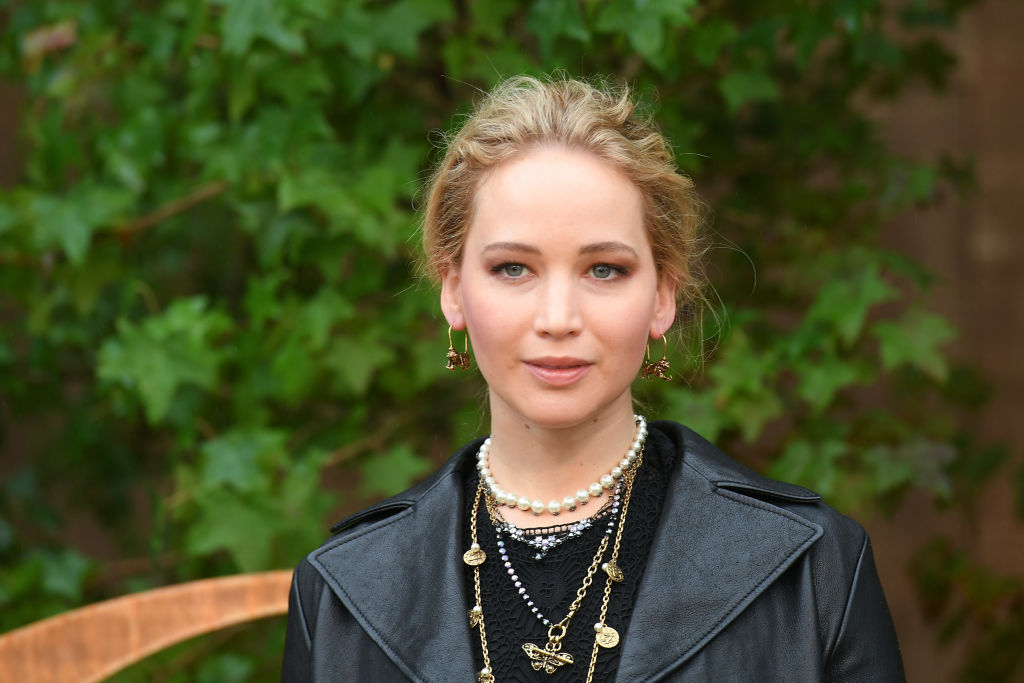 Jennier Lawrence vestida com um look total black sobre um fundo de árvores, cheia de colares e olhando serena para a câmera