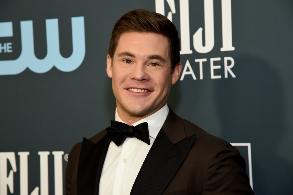 Adam Devine no Critics' Choice Awards em 2020; ele usa um terno preto com gravata borboleta preta e camisa branca; ele está posando e sorrindo para a câmera no tapete vermelho da premiação; a imagem mostra apenas do peito para cima do ator