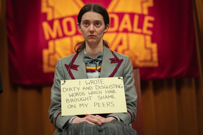 Lily, de Sex Education, está sentada no palco do teatro da escola, na frente de todos os alunos, usando no pescoço uma placa da vergonha, dizendo que ele escreve coisas nojentas