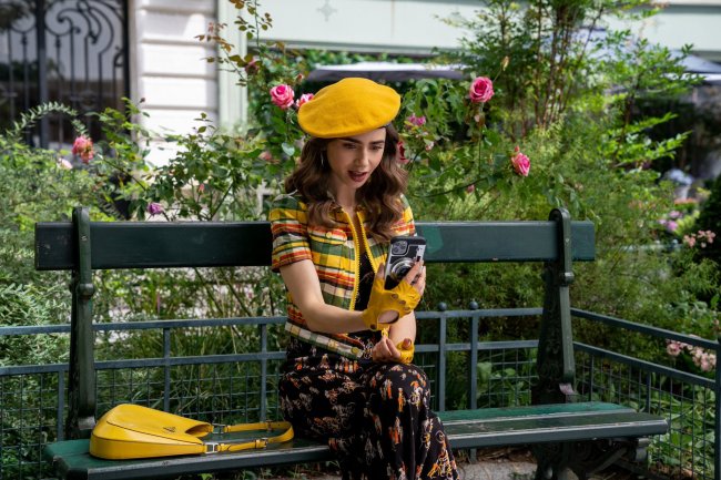 emily em paris segunda temporada Lily Collins como Emily em imagem de da segunda temporada de Emily em Paris; ela está sentada em um banco na rua com paisagem verde com flores rosas ao fundo; ela também está olhando para o celular com expressão chocada e usa um look com detalhes amarelos como boina, luva e a bolsa colocada ao seu lado no banco