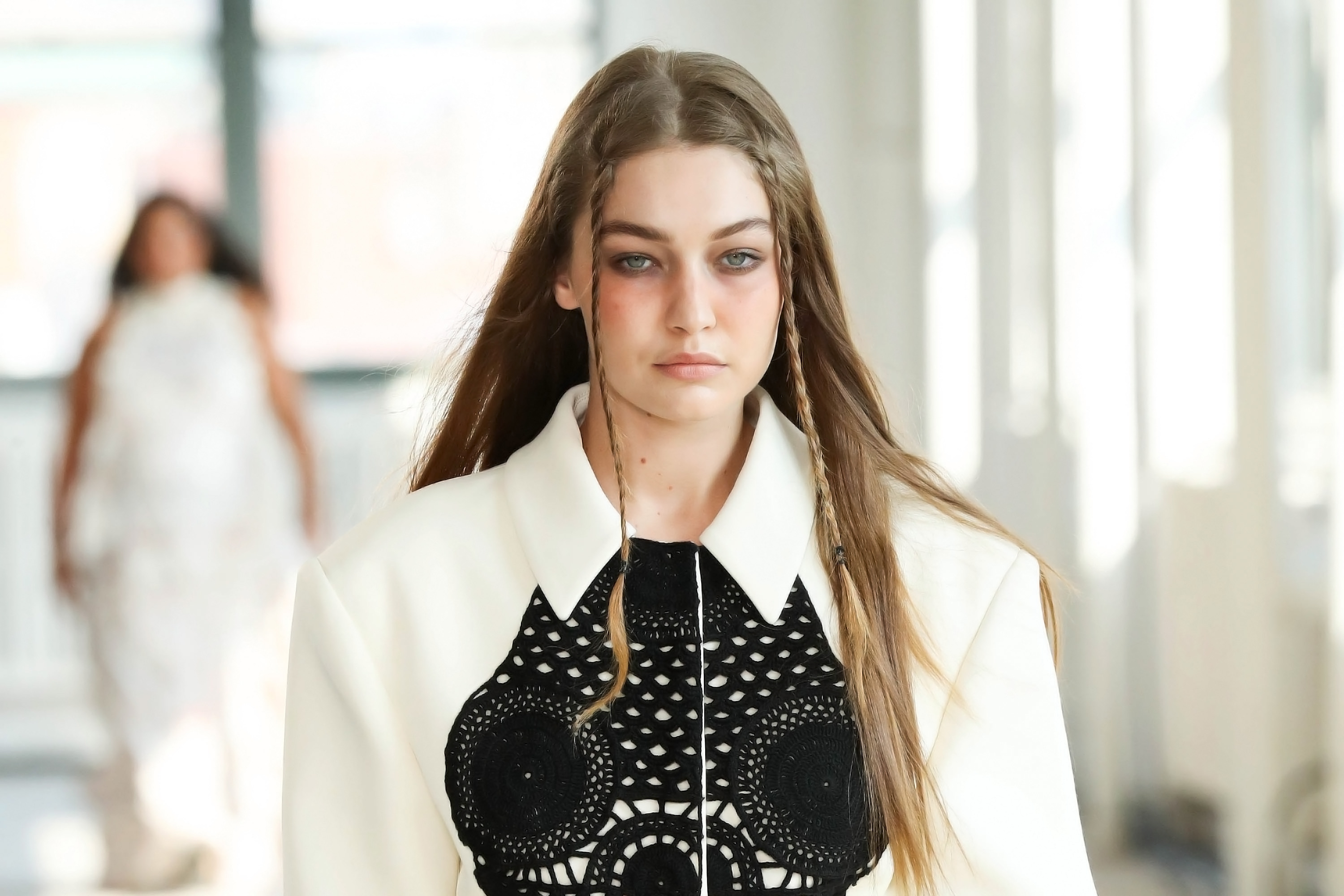 Gigi Hadid no desfile de primavera-verão 2022 da Altuzarra na semana de moda de Nova York. Ela está com expressão facial séria, trancinhas na franja, camisa branca e detalhe preto no decote.