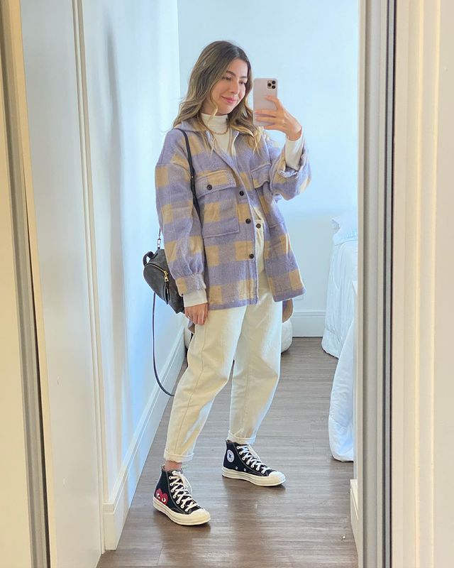 look-all-star-comme-des-garcons Selfie no espelho de uma mulher. Ela usa um casaco xadrez roxo e amarelo, calça branca, tênis preto de cano alto com estampa de coração e mini mochila preta. Ela olha para o celular e sorri levemente.