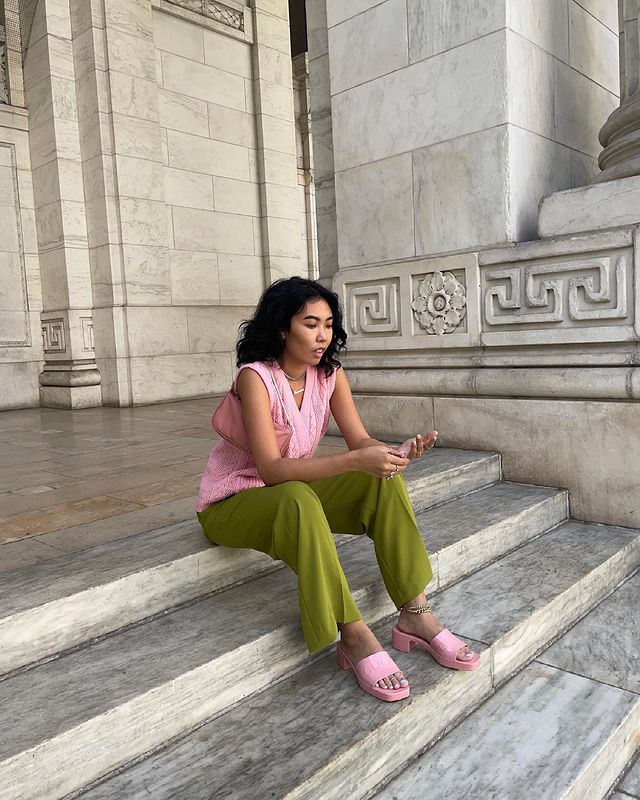 look-colorido-com-tamanco-rosa Foto de uma mulher sentada em uma escada. Ela usa um colete de vovô em um tom de rosa pastel, calça de alfaiataria verde e tamanco rosa pastel. Ela olha para frente e não sorri.