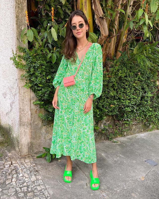 look-com-tamanco-colorido Foto de uma mulher na rua. Ela usa um vestido longo verde, tamanco verde, bolsa rosa, óculos de sol e cabelo solto. Ela olha para a câmera e sorri levemente.