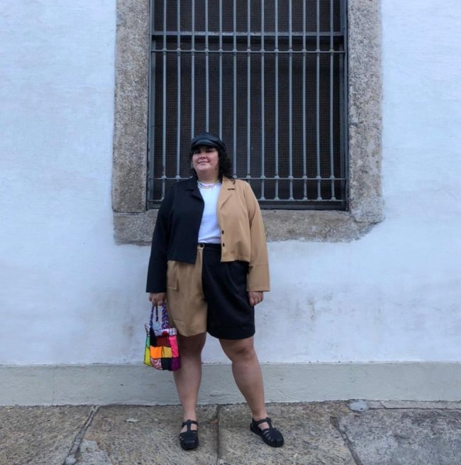 Jovem posando frente a parede branca usando conjunto de peças preta e marrom.