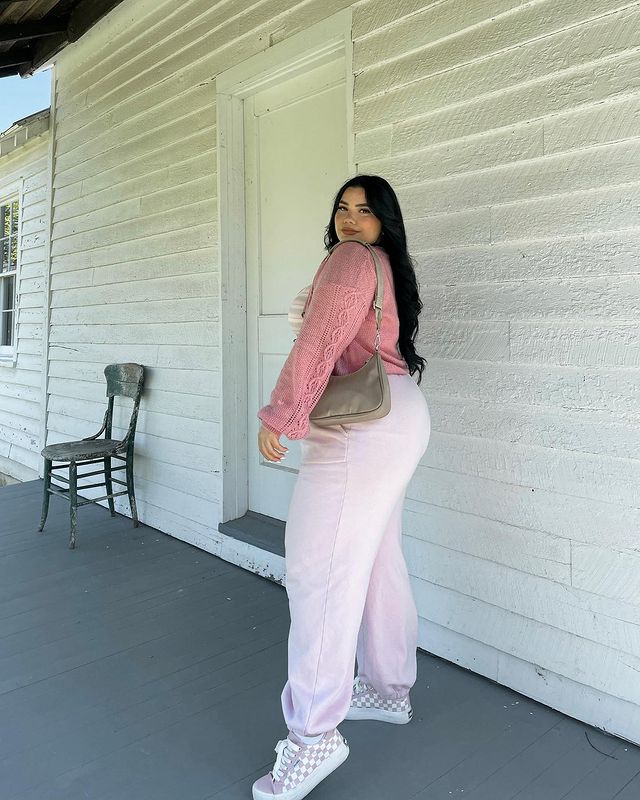 look-tenis-rosa-estampado Foto de uma mulher em um corredor. Ela usa um cardigan rosa, calça moletom rosa e tênis rosa quadriculado. Ela olha para a câmera e sorri levemente.