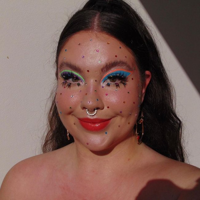 make-artistica-cilios-inferiores-destacos Foto do rosto de uma mulher. Ela usa um penteado meio preso, maquiagem com os olhos coloridos, cílios inferiores destacados com cílios postiços e pedrinhas coloridas em todo o rosto. Ela olha para a foto e sorri.