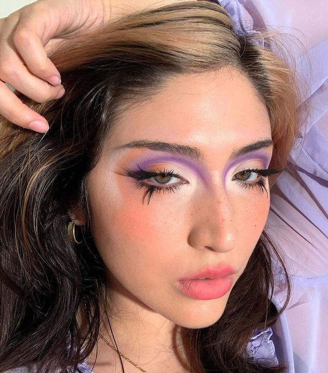 make-artistica-cílios-inferiores Selfie de uma mulher. Ela está com o cabelo solto, maquiagem com sombra colorida, cílio postiço nos cílios inferiores e pele básica. Ela olha para a câmera e não sorri.