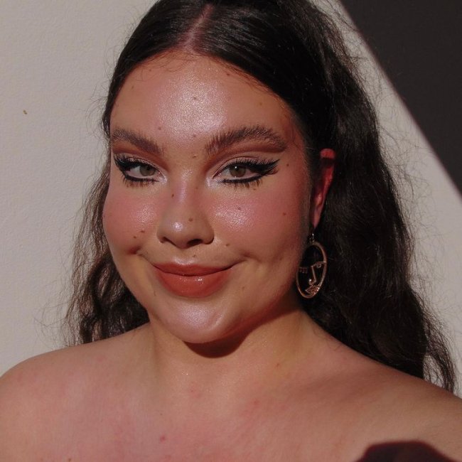 make-cilios-inferiores-destacados Foto de uma mulher. Ela está com o cabelo meio preso em um rabo de cavalo, usa maquiagem com delineado gráfico e cílios inferiores destacados. Ela olha para a câmera e sorri.