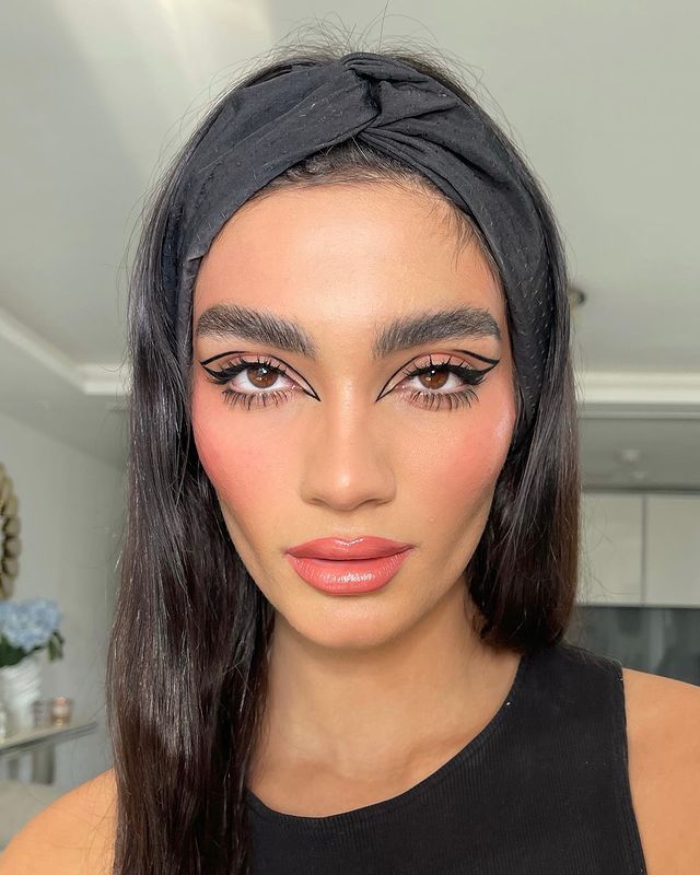 make-com-cilios-inferiores-destacados Foto de uma mulher. Ela usa uma blusa preta, tiara preta no cabelo e está com uma maquiagem básica com um delineador gráfico e cílios inferiores destacados.