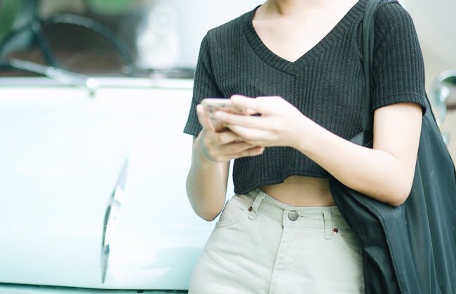 Mulher digitando no celular apoiada no capô de um carro. Não é possível ver o seu rosto.