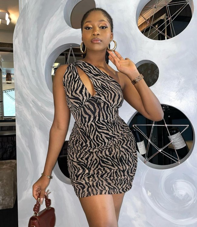 vestido animal print com recorte Jovem posando com uma das mãos apoiadas no brinco, e com expressão séria. Ela segura uma bolsa de mão.