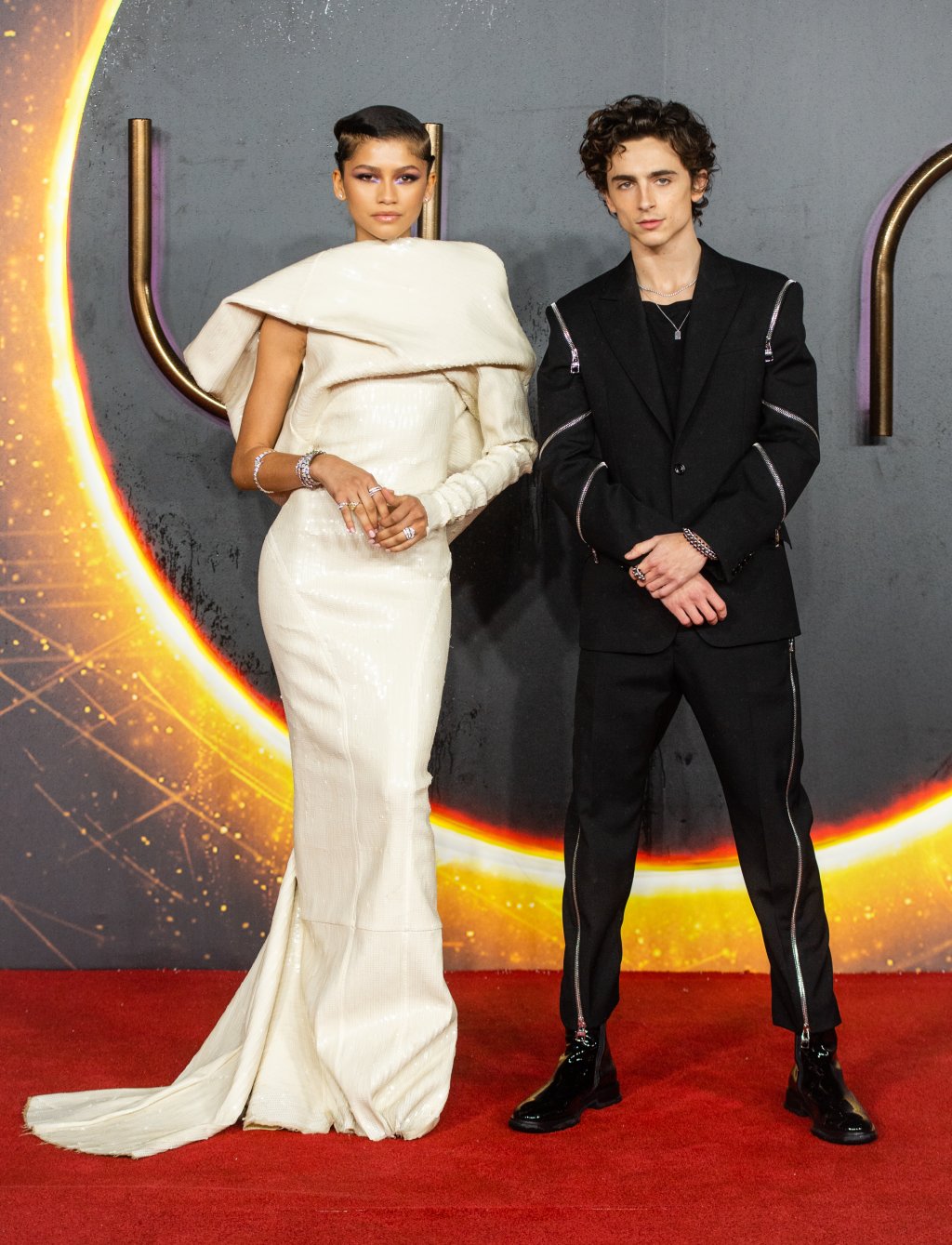 Zendaya e Timothée Chalamet no carpet de Duna; a atriz usa um vestido branco com apenas uma manga comprida enquanto o ator usa um look todo preto com detalhes em dourado; ao fundo é possível ver o logo do filme Duna em um backdrop preto; eles posam com expressões sérias lado a lado com a mãos na frente do corpo em um tapete vermelho