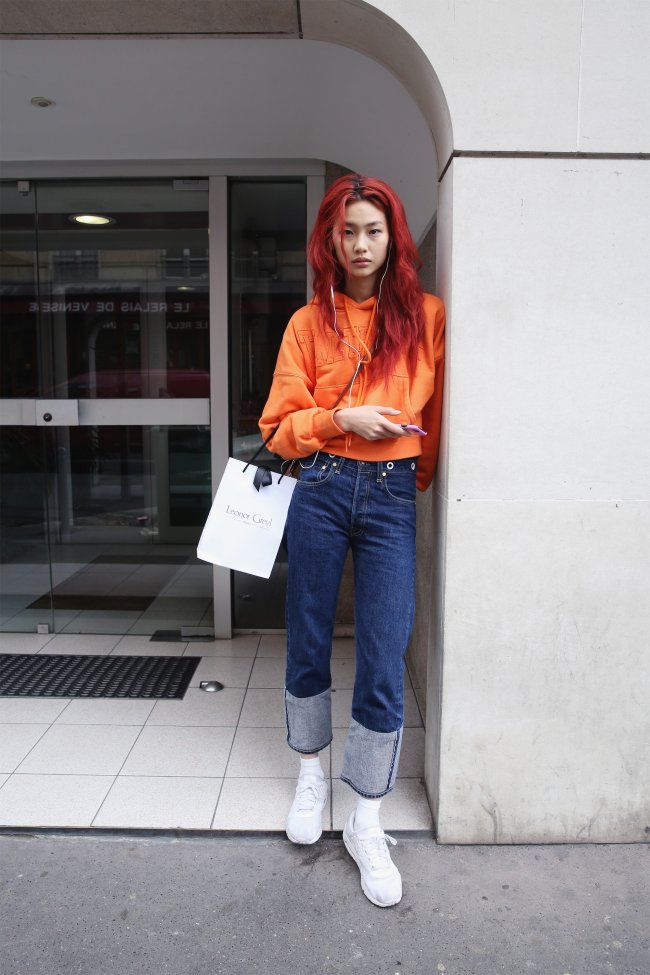 Jung Ho-yeon Jung Ho-yeon encostada na parede usando blusa laranja, calça jeans e com uma expressão séria.