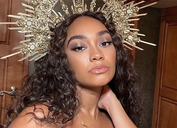 Leigh-Anne-Pinnock de cabelo solto e usando uma coroa dourada na cabeça. Com uma das mãos no pescoço, ela olha para a câmera
