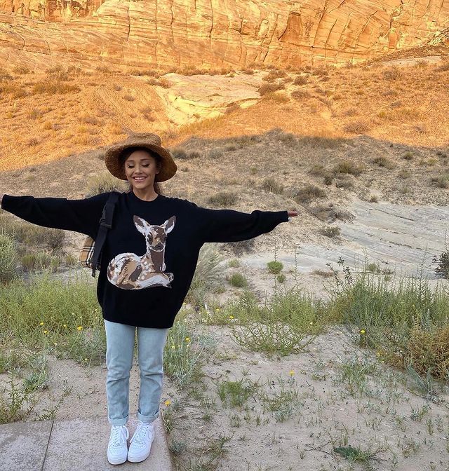 Foto da Ariana Grande em uma paisagem. Ela usa um chapéu marrom, moletom preto, calça jeans, tênis branco e mini mochila. Ela está sorrindo de olhos fechados e com os braços abertos.