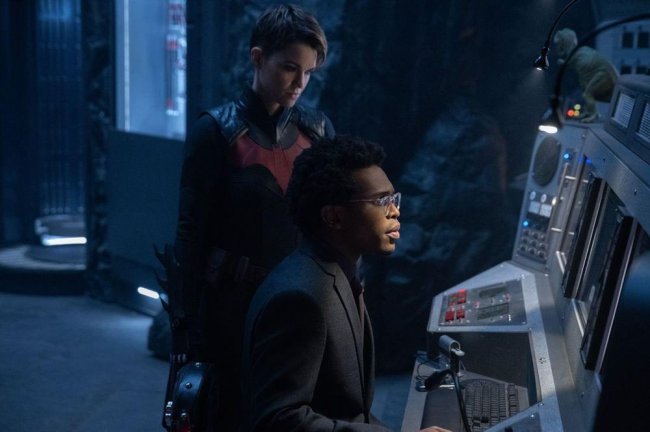 Foto de Ruby Rose e Camrus Johnson em Batwoman. Nela, a atriz aparece em pé com um traje vermelho e preto, enquanto ele está sentado olhando para o computador, de óculos e terno preto.