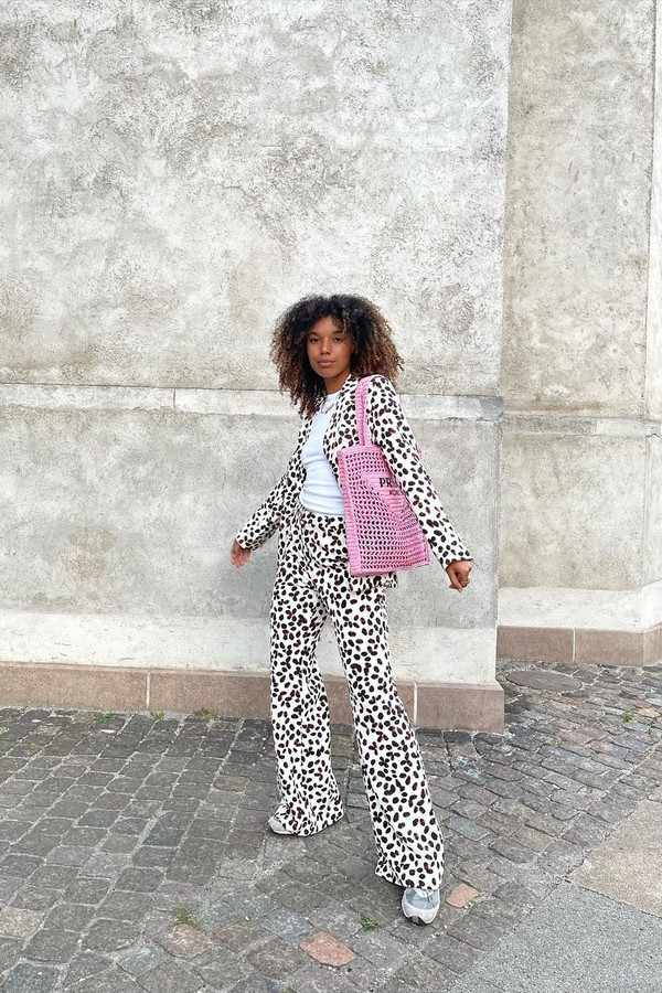 Foto de uma mulher em uma calçada. Ela usa o cabelo solto, camiseta branca, casaco branco com estampa de bolinhas pretas, calça com a mesma estampa, tênis e bolsa rosa de crochê da Prada. Ela olha para a câmera e não sorri.