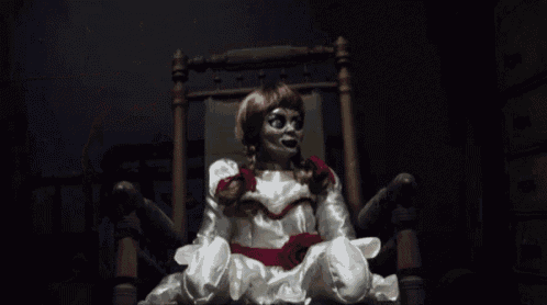 Os 13 bonecos amaldiçoados mais macabros do cinema e da internetboneca_domal Gif da Annabelle. Ela está sentada em uma cadeira de balanço, usa seu icônico vestido branco e vermelho, e olha devagar para a câmera
