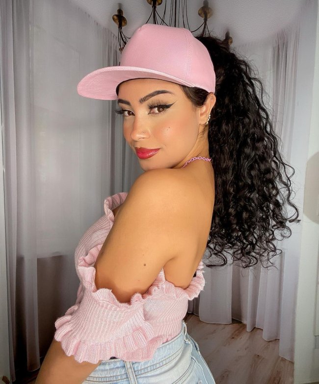 cabelo cacheado com rabo de cavalo Jovem sorridente com boné rosa e blusa da mesma cor. Ela olha de lado para a câmera.