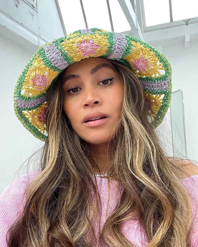 chapeu-de-croche Selfie de uma mulher. Ela usa uma blusa rosa, cabelo solto, maquiagem básica e chapéu de crochê colorido. Ela olha para a câmera e não sorri.