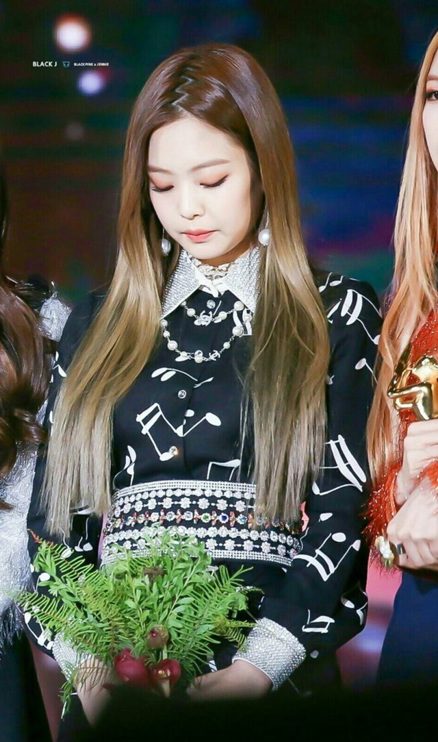 jennie-cabelo Foto da cantora Jennie do BLACKPINK. Ela usa um vestido preto com detalhes brancos e cabelo solto repartido em zigue-zague. Ela olha para baixo e não sorri para a foto.