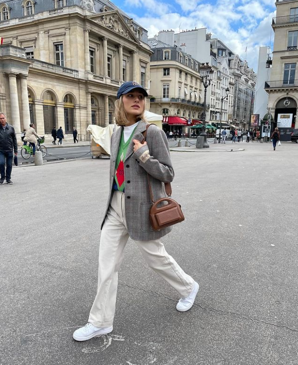 Sasha Meneghel usando boné azul, suéter verde, blazer xadrez, calça off white, tênis branco e uma bolsa marrom em um dos ombros