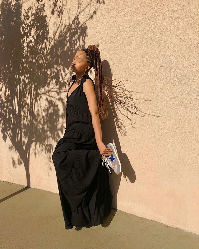 look-vestido-preto-tenis Foto de uma mulher em frente a uma parede rosa. Ela usa um vestido básico longo preto e um tênis esportivo. Ela está segurando o calcanhar esquerdo com a mão esquerda, olha para o lado e não sorri para a foto.