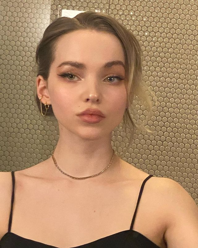 Selfie no espelho da Dove Cameron. Ela usa uma blusa preta de alcinha, colar e brincos dourados, cabelo preso em coque e maquiagem com delineado preto e esfumado marrom. Ela olha para o espelho e não sorri.