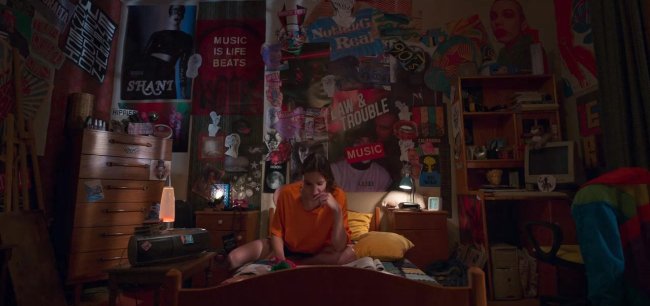 quarto eda love 101 Eda, da série Love 101, estudando sentada na cama com uma camiseta laranja; a iluminação do cômodo vem de um abajur e as paredes estão cobertas por pôsteres e desenhos