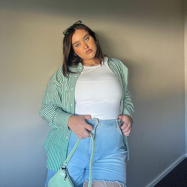 short-jeans-com-camisa Foto de uma mulher encostada em uma parede branca. Ela usa uma camiseta branca, camisa de manga longa verde, short jeans, óculos de sol e bolsa baguete verde. Ela olha para a câmera e não sorri