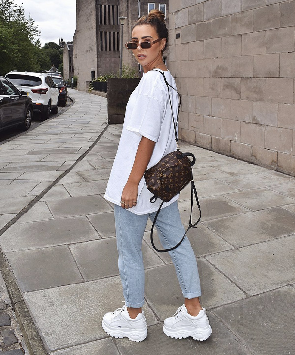 Jovem posando virada de costas com a bolsa pendurada, ela usa calça jeans e tênis branco.