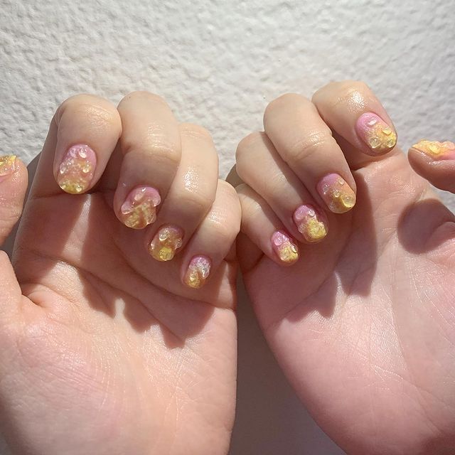 unha-colorida-gota-de-agua Foto de duas mãos com o esmalte degradê de rosa e amarelo com detalhe de gota de água em gel 3D.