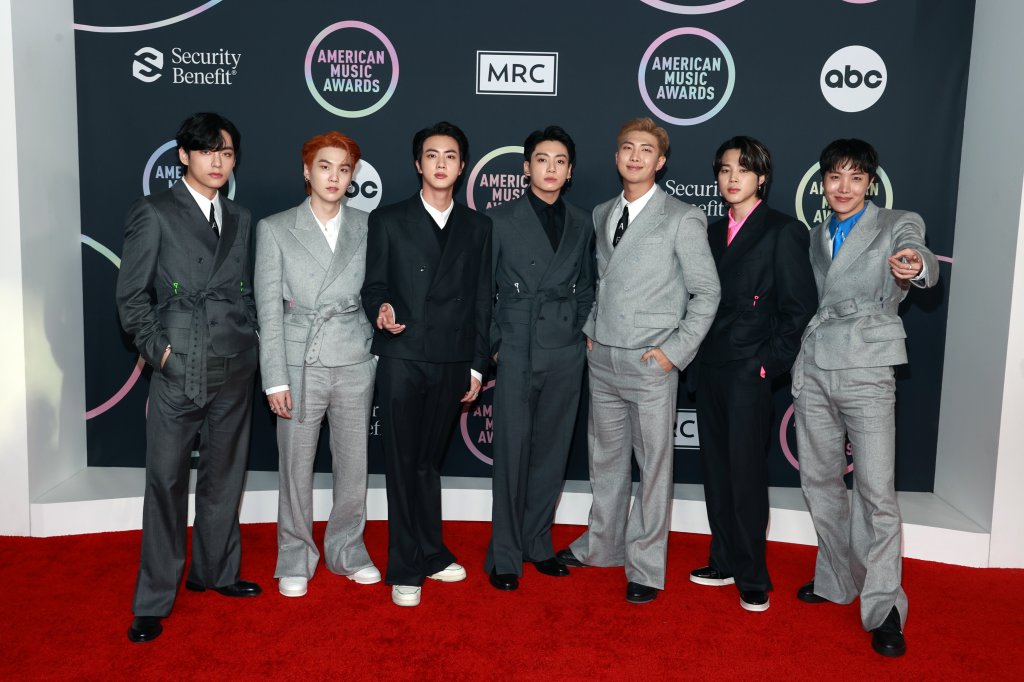 V, Suga, Jin, Jungkook, RM, Jimin, e J-Hope do BTS no American Music Awards 2021 (AMAs); eles estão posando no backdrop da premiação com logos do AMAs, patrocinadores e a emissora ABC; os integrantes usam ternos em tons de cinza e preto e estão com expressões neutras posando no tapete vermelho do evento olhando para câmera com as mãos no bolso