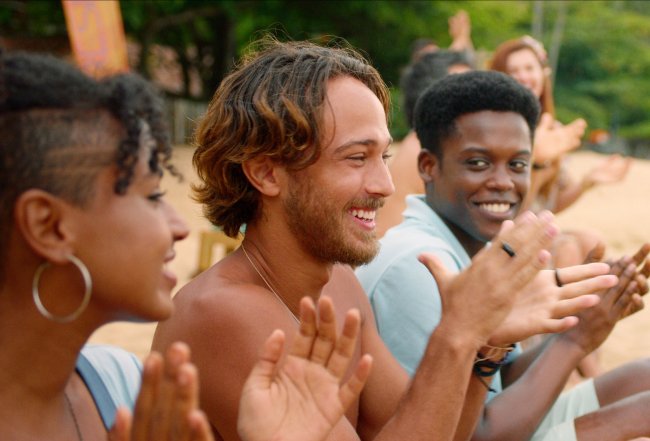 Elenco de Temporada de Verão, a nova série da Netflix; os personagens estão sentados na praia alinhados batendo palmas e sorrindo; a foto foi dirada de perfil e é possível ver areia e folhas de árvores verdes ao fundo