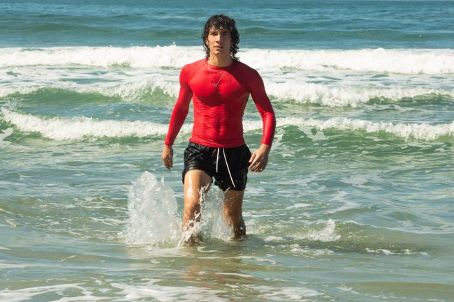 TDV_107-Diego_0084R Imagem de divulgação de cena com Jorge Lopez como Diego em Temporada de Verão, nova série da Netflix; ele está saindo do mar com a roupa molhada e veste uma bermuda preta com uma camisa vermelha de manga comprida; ao fundo é possível ver o mar e a espuma de algumas ondas