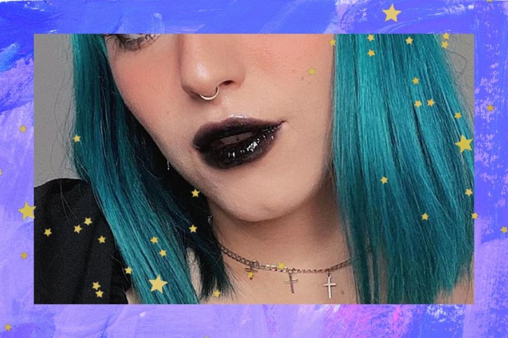 Montagem com o fundo roxo com a foto do close do rosto de uma mulher com detalhe de estrelinhas douradas nas bordas. A mulher está com o cabelo azul, curto e liso solto, usa colar com pingentes de cruz e batom preto e gloss.