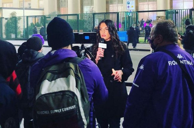 Foto de Greta Beccaglia falando durante transmissão ao vivo, rodeada de torcedores do Fiorentina. Ela é branca, tem o cabelo preto e usa um sobretudo preto