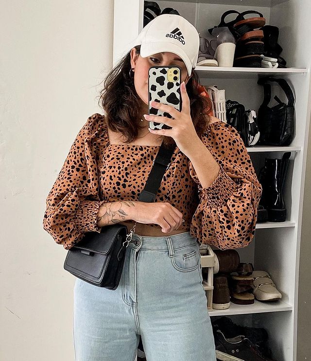 look-animal-print-com-bone Selfie de uma mulher no espelho. Ela usa uma calça mom jeans, blusa com manga bufante com estampa de animal, boné com aba curva branco e bolsa preta. Ela olha para o celular e não sorri.