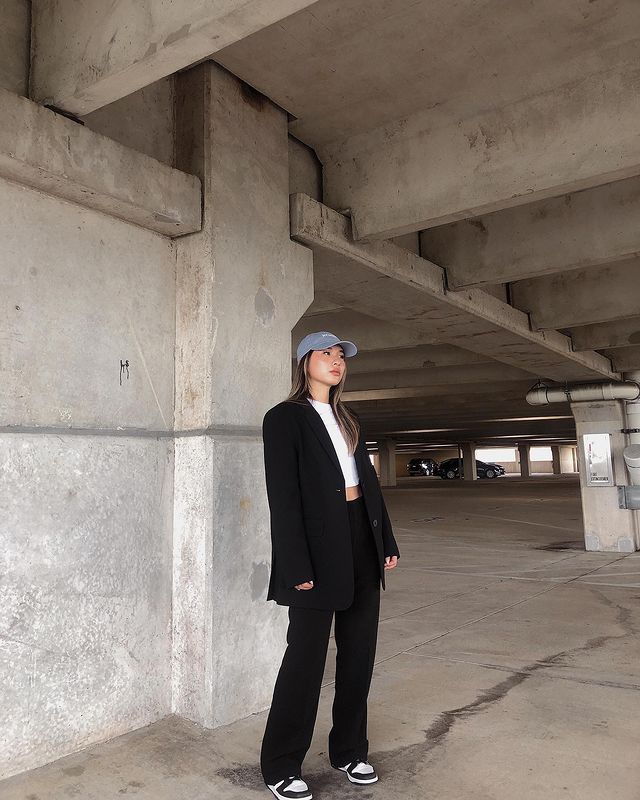 look-blazer-com-dad-cap Foto de uma mulher em uma garagem de carros. Ela usa uma blusa branca, terno preto, calça de alfaiataria preta e boné com a aba curva azul. Ela olha para frente e não sorri.