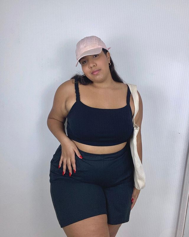 look-com-bone-de-pai Foto de uma mulher em frente a uma parede. Ela usa top cropped preto, bermuda preta, ecobag e boné com a aba curva rosa. Ela olha para a câmera e não sorri.