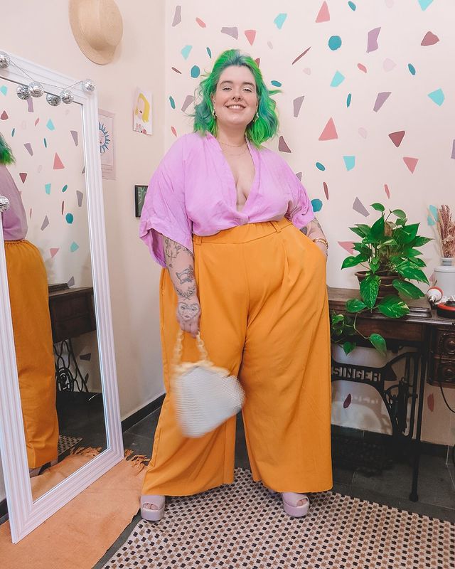 look-com-calca-laranja Foto de uma mulher em um quarto. Ela usa uma camisa lilás, calça laranja, tamanco rosa e bolsa branca. Ela está com o cabelo curto e verde solto, olha para a câmera e sorri.
