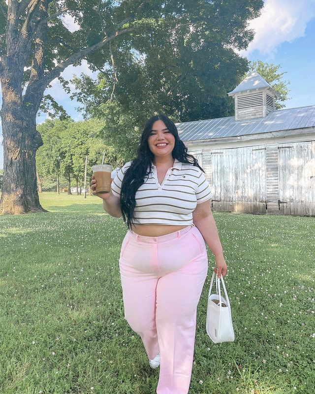 Foto de uma mulher em um lugar com árvores e grama. Ela usa um top cropped branco com listras marrom, calça rosa clara e bolsa branca. Ela está com o cabelo solto, segura um copo com a mão direita e a bolsa com a mão esquerda, olha para a câmera e sorri.