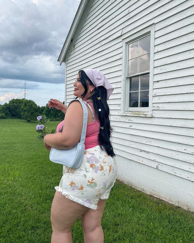 look-lenco-no-cabelo Foto de uma mulher em frente a uma casa. Ela usa uma regata rosa, short estampado com flores, bolsa baguete azul e lenço rosa como acessório no cabelo. Ela está de lado, olha para o lado e sorri levemente.