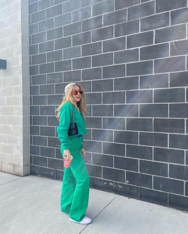 look-monocromatico-verde Foto de uma mulher em uma calçada. Ela usa um moletom verde, calça verde, tênis branco, lenço colorido no cabelo, bolsa baguete preta e óculos de sol com armação vermelha. Ela segura um copo na mão direita, olha para a frente e sorri levemente.