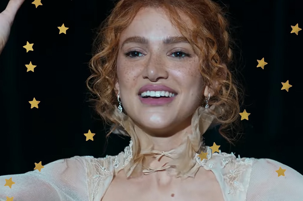 Manu Gavassi no trailer do álbum visual GRACINHA; ela está sorrindo com o cabelo preso e a franja solta; ela está com uma roupa de renda branca e os braços levantados em um palco; o fundo está escuro e um holofote de luz branca ilumina o rosto da cantora; estrelas amarelas decoram a imagem